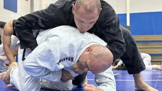 Older BJJ white belt in a gi defending as a black gi partner pressures from the top during class