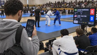 Blue belt competitor checking the fight order on his phone at a BJJ tournament while two blue belts grip fight next to a scoreboard