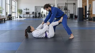 Woman in a white gi on her back using open guard, gripping the sleeves of a standing partner with a foot on his hip to keep distance