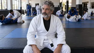 Grey-bearded man in his fifties wearing a new white gi and white belt, sitting on the mat edge before his first BJJ class